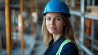 Young Caucasian female construction engineer wearing blue hard hat and safety vest smiles confidently at construction site, representing women in engineering and building industry.