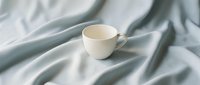 Empty white ceramic cup on soft gray fabric with natural folds and wrinkles, suitable for beverage mockups and product presentations.