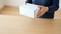 Man in dark navy sweater holding white cardboard box on wooden table. Suitable for shipping, delivery, unboxing, e-commerce, and logistics concepts.