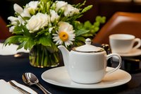 Elegant white teapot and cup set on formal dining table with fresh floral centerpiece featuring roses and daisies, creating luxurious afternoon tea atmosphere.