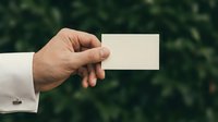 Male hand in white suit holding blank business card outdoors, green bokeh background, copy space for branding and corporate identity.