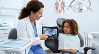 Female dentist showing dental X-ray results on tablet to young African American girl patient in modern clinic examination room with medical equipment.