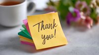 Handwritten thank you message on yellow sticky note with colorful paper stack, cup of tea and pink flowers in soft focus background, expressing gratitude concept.