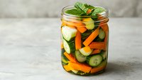 Fresh pickled vegetables in glass jar with cucumber slices, carrot sticks, bell peppers and herbs on light background. Homemade fermented food concept.