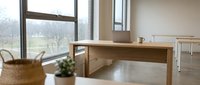 Minimalist wooden desk in bright office space with large windows overlooking nature, potted plant, and wicker basket for storage and workspace organization.
