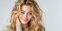 Young Caucasian woman with tousled blonde wavy hair smiling gently while wearing a beige knit sweater against neutral gray background.