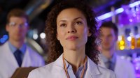 Young female medical professional with curly hair in white lab coat looking up thoughtfully in modern laboratory setting with purple lighting and colleagues in background.