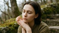 Young woman with dark hair in contemplative pose outdoors, hands clasped near face, peaceful expression in natural forest setting with soft lighting.