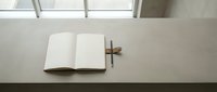 Minimalist workspace with open blank notebook and wooden cross on white desk near window for spiritual writing, journaling, prayer planning, and faith-based content creation.