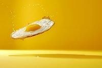 Levitating fried egg with crispy edges and bright yellow yolk on vibrant yellow background with splashing oil drops, minimalist food photography composition.