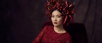 Young woman with East Asian ethnicity wearing elaborate red textured garment and matching ornamental headdress with flowing elements against dark backdrop. Suitable for fashion, beauty, editorial, and artistic projects.