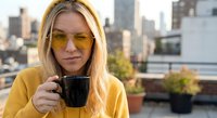Young blonde woman in yellow sunglasses and hoodie drinking coffee on urban rooftop terrace with city skyline background during golden hour morning routine.
