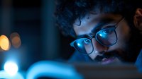 Young bearded Middle Eastern man in glasses illuminated by blue computer screen light at night, focused expression while working late with bokeh background effects.