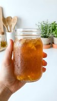 Hand holding a glass jar filled with cold iced tea or cola beverage with condensation droplets, kitchen setting with wooden utensils and potted herbs.