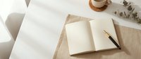 Open blank notebook with pen on linen cloth, coffee cup and eucalyptus branch on white desk in soft natural light.