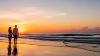 Romantic couple silhouettes walking on beach at golden sunset with ocean waves reflecting warm light on wet sand creating peaceful evening atmosphere.