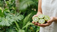 Woman in light linen shirt holding fresh kiwi fruit slices in hands outdoors among green foliage, promoting healthy eating and organic produce.