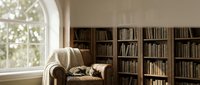 Cozy reading nook with sleeping cat on armchair near window and wooden bookshelf in warm sunlit home library interior for relaxation and literature lovers.