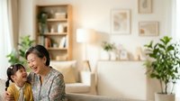 Mature Asian woman and young daughter laughing together in bright living room with plants, shelving, and warm lighting, representing family bonding and home comfort.