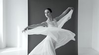Graceful female ballet dancer in flowing white dress performing elegant movement in modern studio with geometric architectural background.