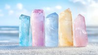 Colorful healing crystal points arranged on beach sand against blurred ocean background, featuring pastel blue, pink, yellow and rose quartz minerals in natural light.