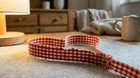 Red checkered gingham ribbon on white surface in cozy home interior with warm lighting and rustic wooden furniture background for crafting projects.