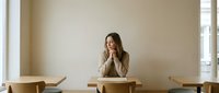 Young woman sitting alone at wooden table in minimalist cafe interior, contemplating with hand on chin in thoughtful pose for lifestyle and business concepts.