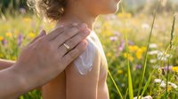 Adult applying sunscreen lotion to child's shoulder in sunny meadow with wildflowers and grass for skincare and sun protection.
