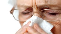 Senior woman with glasses wiping nose with tissue, showing facial expression of discomfort during cold or allergy symptoms, close-up view of face and hands.