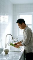 Asian man washing fresh asparagus spears under running water in modern white tiled kitchen. Healthy cooking and meal preparation at home.