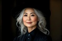 Elegant mature Asian woman with silver gray wavy hair wearing black hat and shirt, confident smile against dark background. Professional portrait with dramatic lighting.