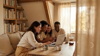 Family of four, mixed ethnicities, gathered around laptop at wooden table in bright living room with bookshelves and natural light, suitable for web design, education, and lifestyle content.