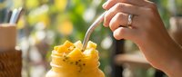 Closeup of woman's hand with engagement ring stirring yellow passion fruit smoothie with metal straw outdoors, summer beverage concept.