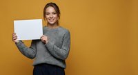 Young Caucasian woman in gray sweater holding blank white paper sheet against yellow background, smiling at camera for business presentation mockup.