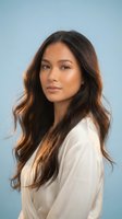Young Asian woman with long wavy brown hair wearing white blazer against light blue background for professional headshot and business portrait.