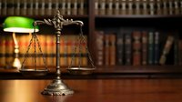 Brass scales of justice on wooden desk in law library with green banker lamp and legal books creating professional attorney office atmosphere.