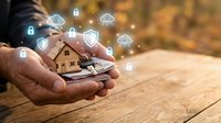 Hands holding miniature house model with digital security icons and cloud symbols floating above representing smart home technology and property protection.