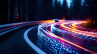 Vibrant light trails curve along dark mountain road at night, creating colorful streaks of red and blue against forest backdrop. Long exposure photography captures motion.