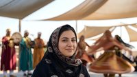 Young Middle Eastern woman wearing traditional black headscarf smiling at camera in bustling marketplace with colorful fabrics and textiles in background.