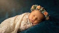 Peaceful newborn baby girl sleeping wrapped in delicate lace blanket wearing floral crown headband for maternity photography and birth announcements.