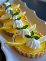 Mini lemon tarts with whipped cream rosettes and fresh mint garnish on white serving platter, bright yellow curd filling in crisp pastry shells for dessert menu.