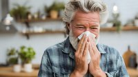 Senior Caucasian man with gray hair and beard blowing nose into tissue, suffering from cold or allergies in cozy kitchen with plants in background.