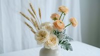 Elegant floral arrangement with peach ranunculus flowers, wheat stalks and dusty miller foliage in glass vase on white table for home decor and styling.