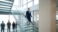Professional businesswoman in navy suit walking down modern glass staircase in contemporary corporate office building with colleagues in background.