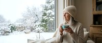 Young blonde Caucasian woman in cozy beige sweater and knit hat enjoying hot beverage by window overlooking snowy winter landscape for seasonal lifestyle content.