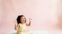Young girl with curly dark hair wearing yellow dress, sitting on white surface against soft pink background, hands raised in joyful gesture.
