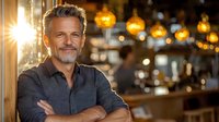 Confident mature Caucasian man with gray hair and beard smiling in restaurant interior with warm lighting and bokeh background, arms crossed in casual attire.