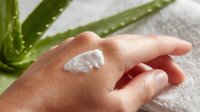 White moisturizing cream on female hand with aloe vera plant in background on white towel, demonstrating skincare and beauty product application for cosmetic advertising.