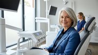 Senior Caucasian woman smiling confidently in modern dental office chair, professional healthcare setting with advanced equipment and staff in background.
