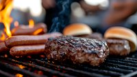 Juicy beef patties and hot dogs grilling on barbecue with flames and smoke. Close-up of meat cooking on grill grates shows charred texture and sizzling fat.